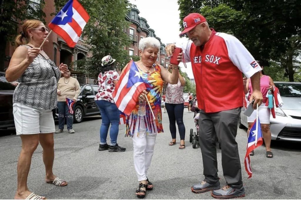 photo of people at a Puerto Rican festival - a woman holding a Puerto Rican flag watches an older couple starting to dance together (they are also holding Puerto Rican flags)