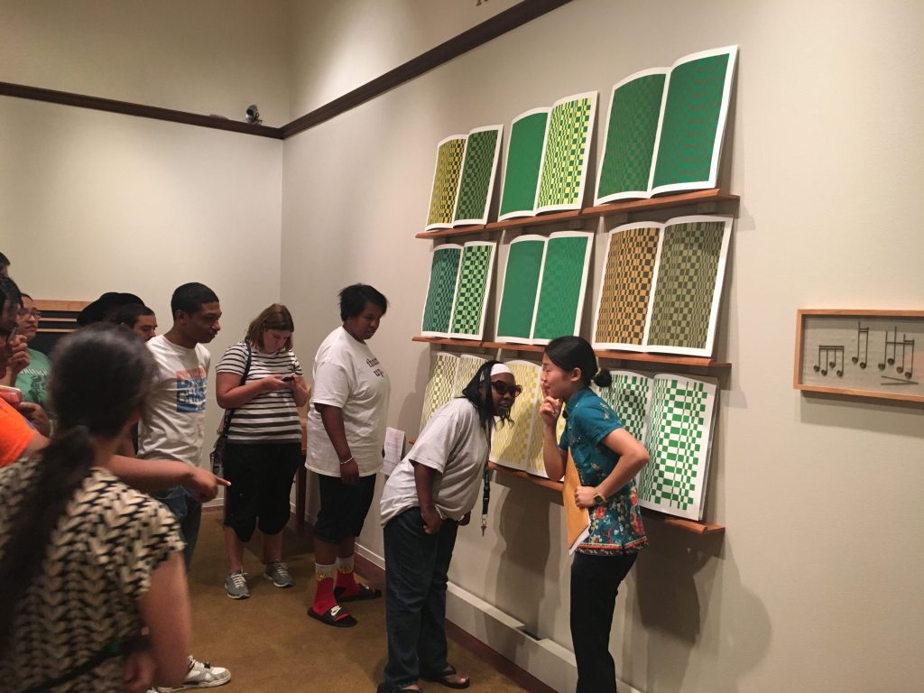 photo of exhibit goers standing before a wall with 3 shallow shelves, graphic prints in greens and browns on the shelves