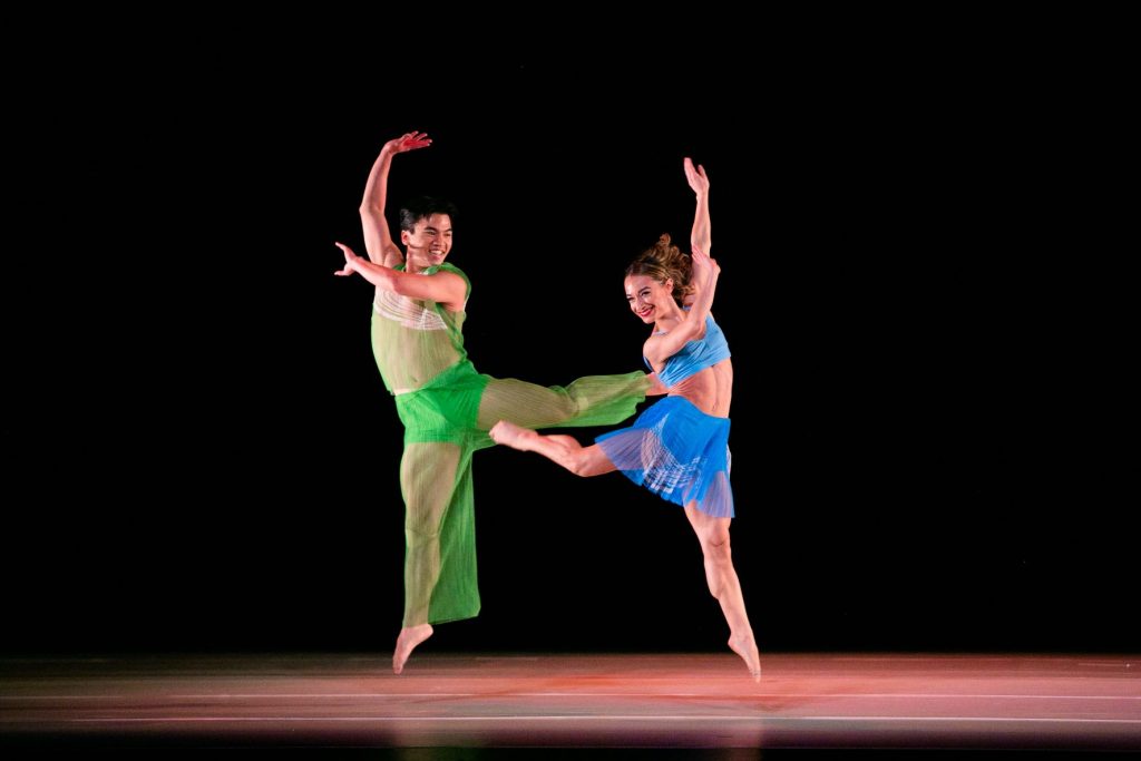 photo of 2 smiling dancers on a stage, both mirroring each other's movements - one arm is raised and the other is crossing the body and one leg extended and bent behind them.