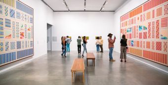 photo of a gallery at the Institute of Contemporary Art - a handful of visitors standing in the back ground, a wooden bench cuts through into the foreground. Large canvases hang of walls to teh left anf right side of the frame