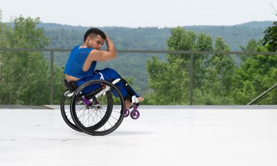 Three disabled and non-disabled dancers perform on a white outdoor stage, which opens up to a majestic background of trees, rolling hills and sky. In energetic unison, the three dancers curl into a ball, with lower limbs raised and upper limbs wrapped protectively around their heads. David and Zara balance on their tailbones, and Janpi balances in a one-handed wheelie in their wheelchair. All three dancers wear royal blue knit cropped shirts and matching pants.