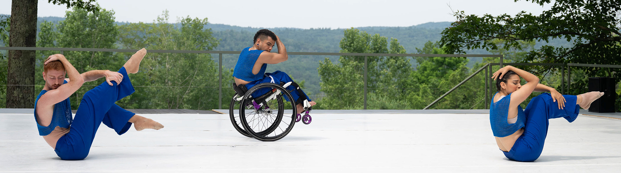 Three disabled and non-disabled dancers perform on a white outdoor stage, which opens up to a majestic background of trees, rolling hills and sky. In energetic unison, the three dancers curl into a ball, with lower limbs raised and upper limbs wrapped protectively around their heads. David and Zara balance on their tailbones, and Janpi balances in a one-handed wheelie in their wheelchair. All three dancers wear royal blue knit cropped shirts and matching pants.