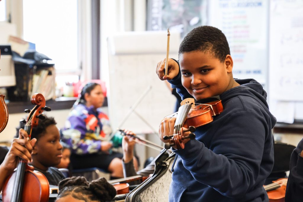 photo of a young man looking at the camera while holding a violin, the neck also pointing at the camera. His chin is on the instrument and he's smiling, the bow poised in the air to begin playing