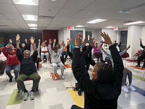 photo of a room of seated older adults all raising their arms while participating in a movement class