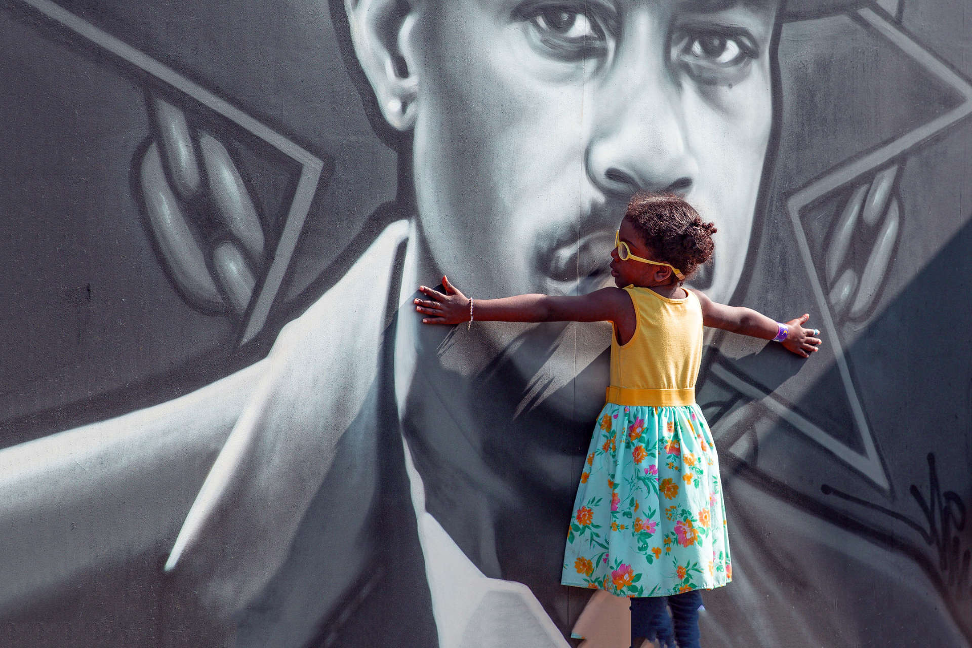photo of a black girl in yellow glasses and a yellow dress holding her arms open to embrace a black and white painted mural depicting a black man's face