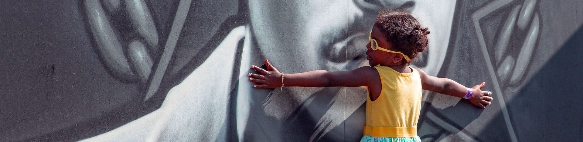 photo of a black girl in yellow glasses and a yellow dress holding her arms open to embrace a black and white painted mural depicting a black man's face