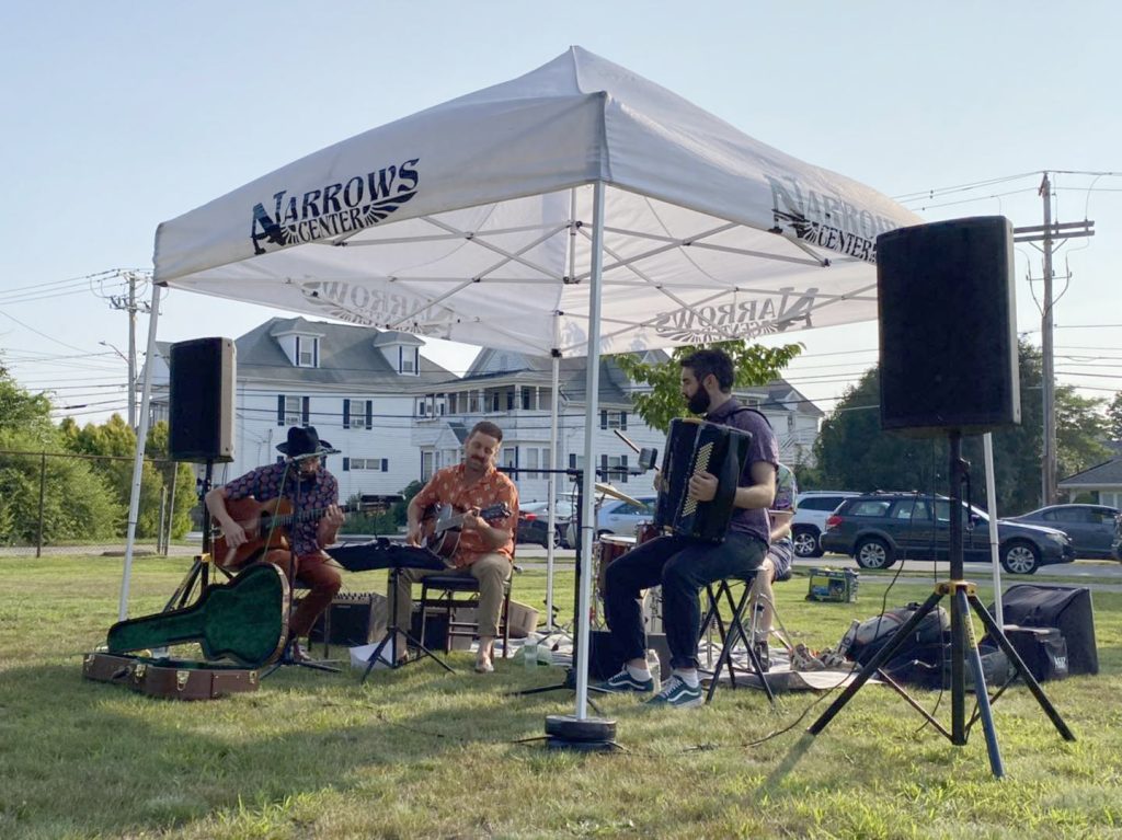 photo of a trio of musicians seated under a white tent playing on a grass commons