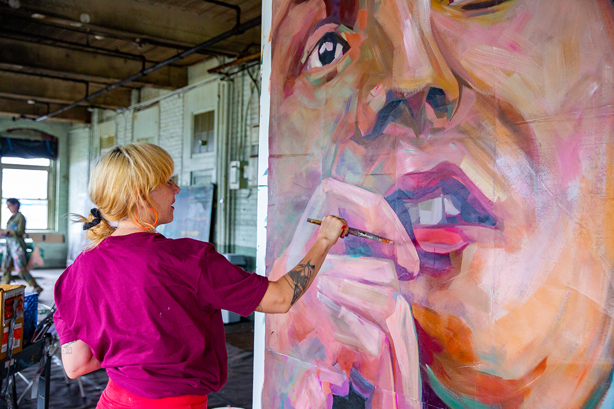 photo of a painter holding a brush, working on a large painting of a person's face