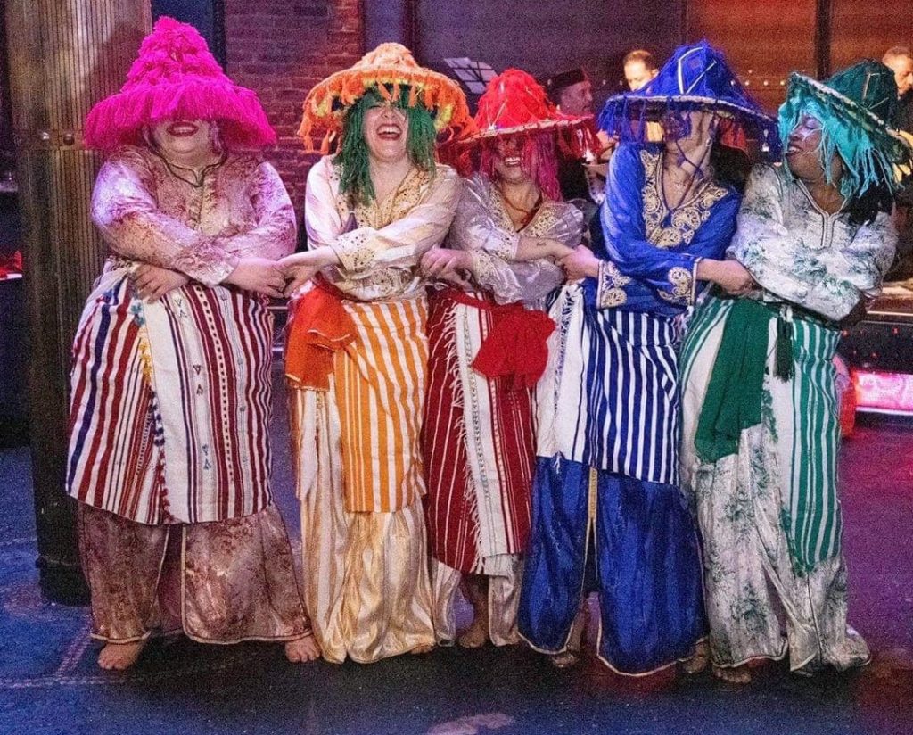 photo of 5 women holding hands with crossed arms and laughing. Each is dressed in a different monochrome costume and hat with long fringe dangling from the wide brims, covering part of their faces