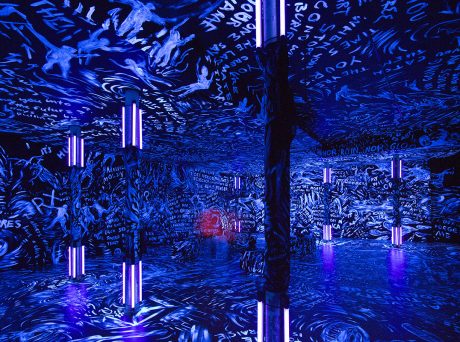 photo of a darkened gallery covered in blue paint with words and figures painted in light colors covering every surface. Blacklights on 3 columns make the lighter paint glow