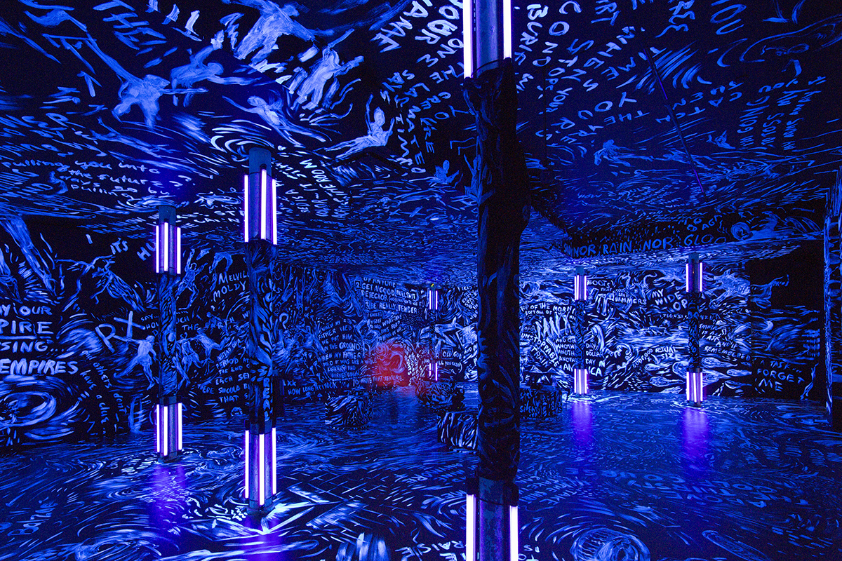 photo of a darkened gallery covered in blue paint with words and figures painted in light colors covering every surface. Blacklights on 3 columns make the lighter paint glow 