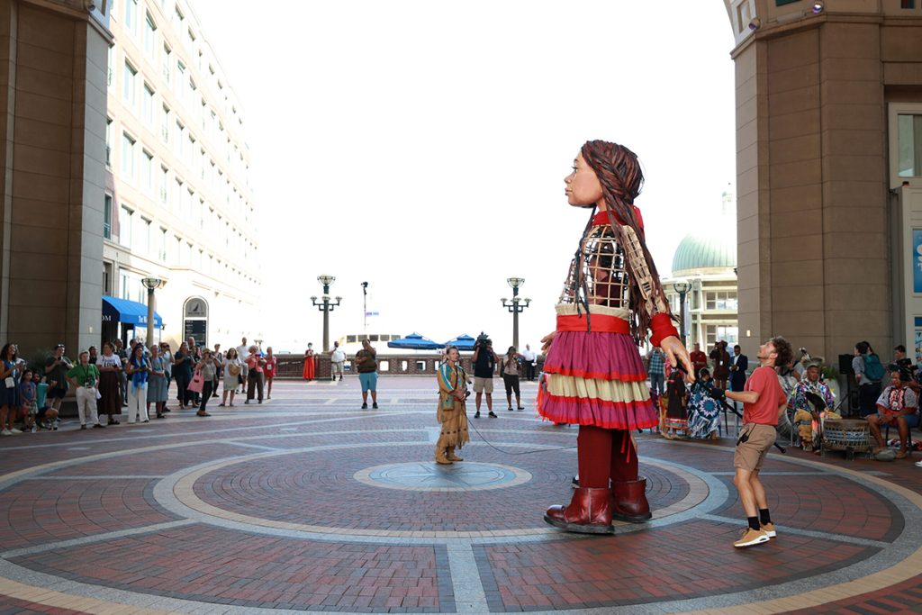 photo of puppeteers manipulating Little Amal, a 10 foot tall puppet, along Boston's Rowes Wharf as onlookers watch