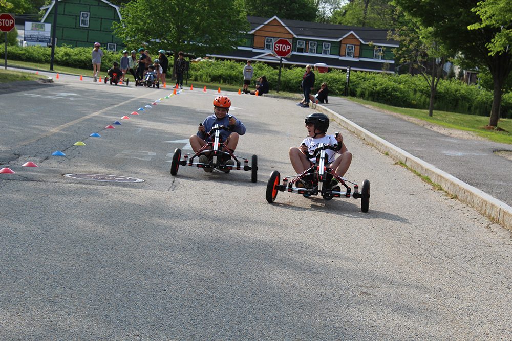 photo of two students wearing helmets riding go-carts down a paved road. One is a bit farther ahead, his head turned back to look at the 2nd go cart