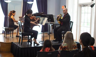 photo of 3 musicians (pianist, violinist and french horn) performing on a small platform stage before a handful of seated audience members