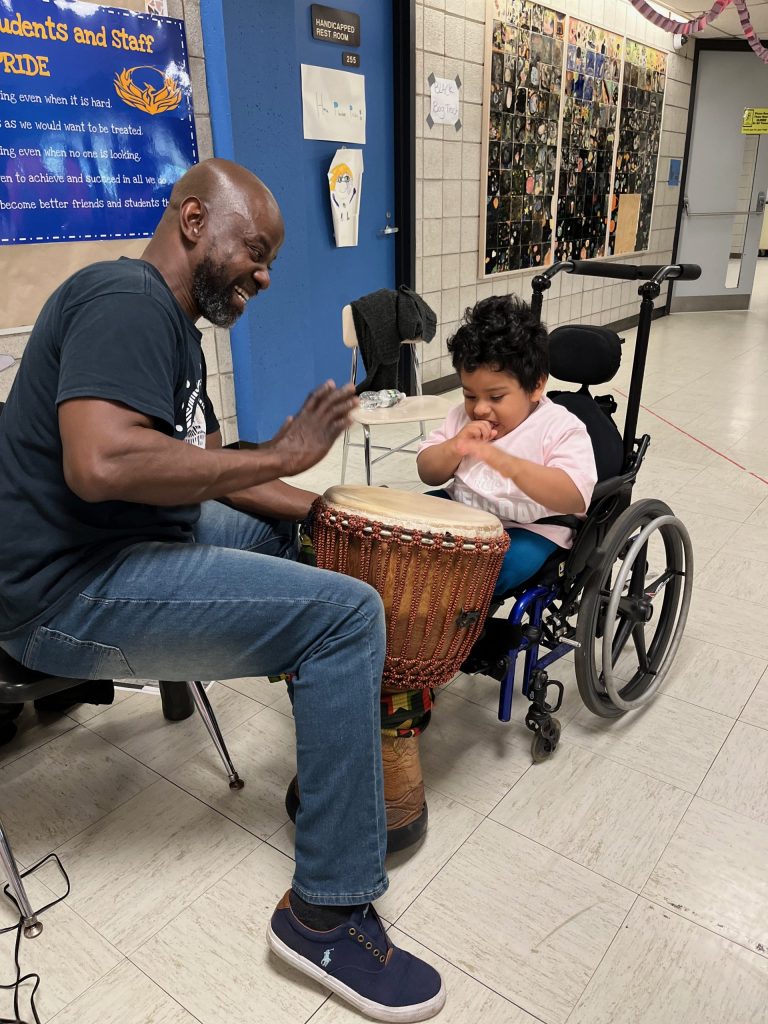 An adult seated in a chair hits an African drum across from a student seated in a wheelchair hitting the same drum