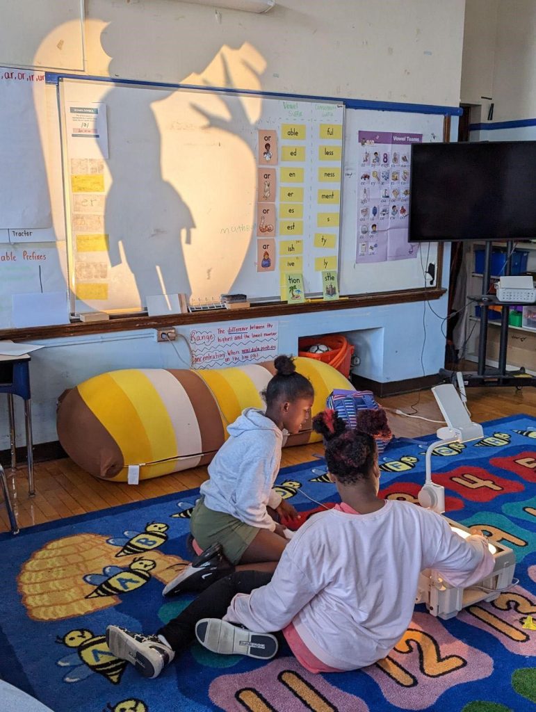 Two students sitting on a colorful classroom rug looking at an overhead projector as they put on a shadow puppet show which is projecting on a whiteboard
