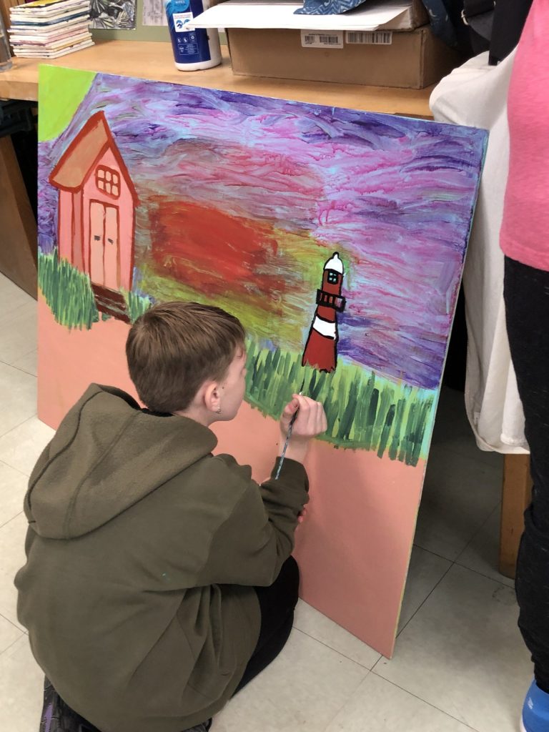 Student crouching with a paintbrush in hand, the wood panel in front of them shows an in-progress lighthouse and seascape 