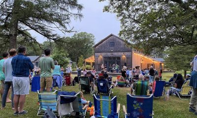 photo of an out door music performance with numerous lawn chairs in the foreground and some people standing facing away to the stage in the background