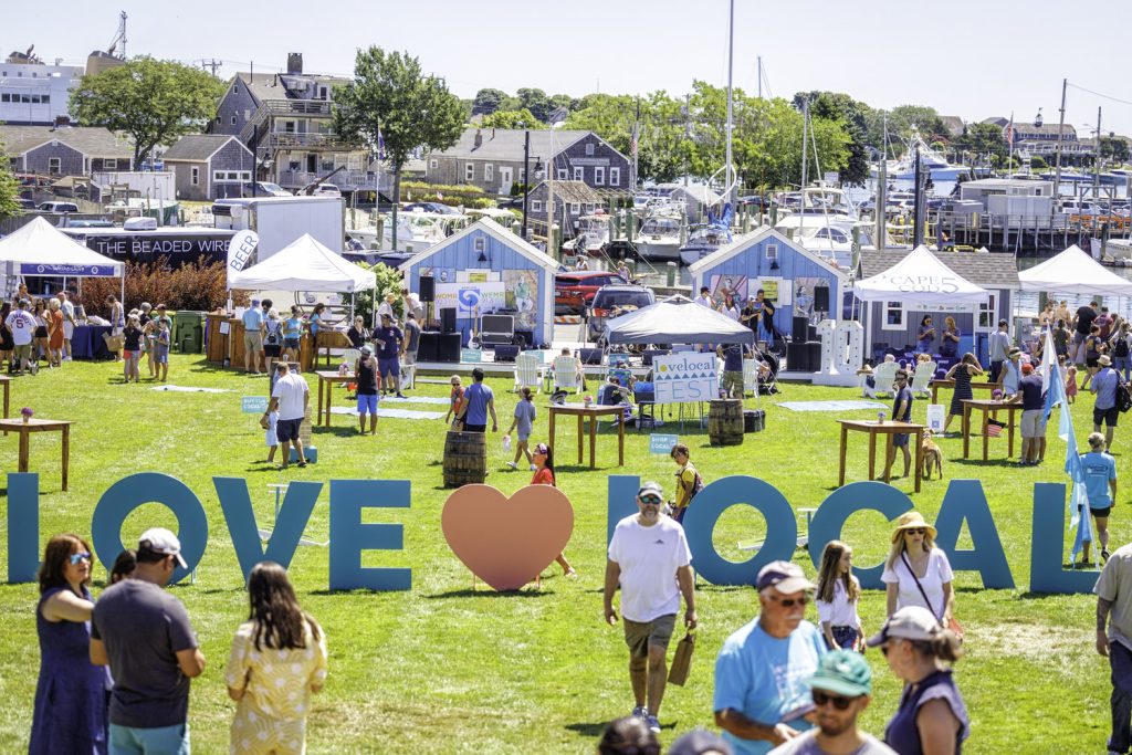 photo of an outdoor festival, people walking in a green space that is peppered with white tents, in the back ground you can see ship masts. In the foreground an art installation with the letters spelling "Love Local" stand amidst festival-goers