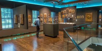 A person stands alone in a museum gallery, viewing historical artifacts in a glass enclosure, the wood floors gleam warmly thanks to the robin's egg blue accented light along the ceiling