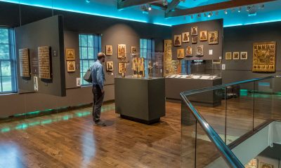 A person stands alone in a museum gallery, viewing historical artifacts in a glass enclosure, the wood floors gleam warmly thanks to the robin's egg blue accented light along the ceiling