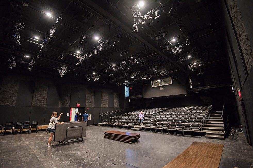 photo of the inside of a black box theater with the pov from the stage. Rows of empty chairs fill the mid and background. 2 theater works roll folding tables across the stage towards the chairs