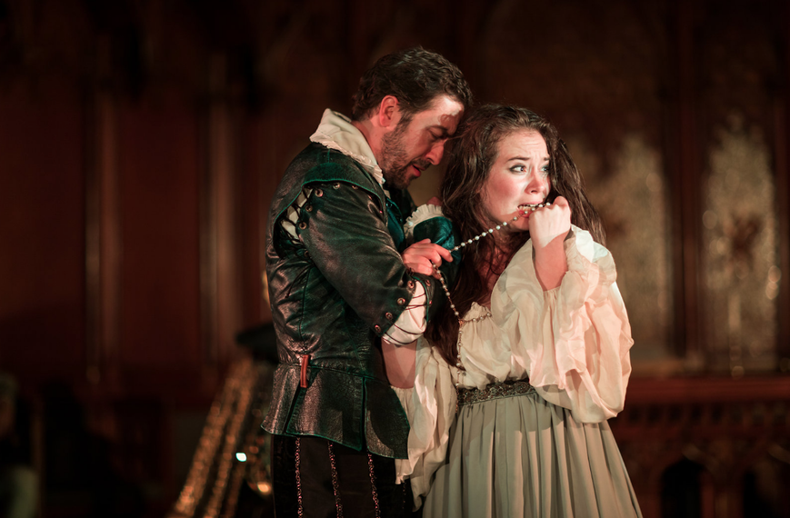 photo from a theater performance of Hamlet. A man and woman embrace. He's looking worried, his eyes cast down. She looks worried, too. Has her string of pearls in her mouth while looking off stage right.