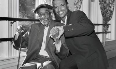 black and white photo of an older Black man sitting with a younger Black man crouching behind him, and holding his hand