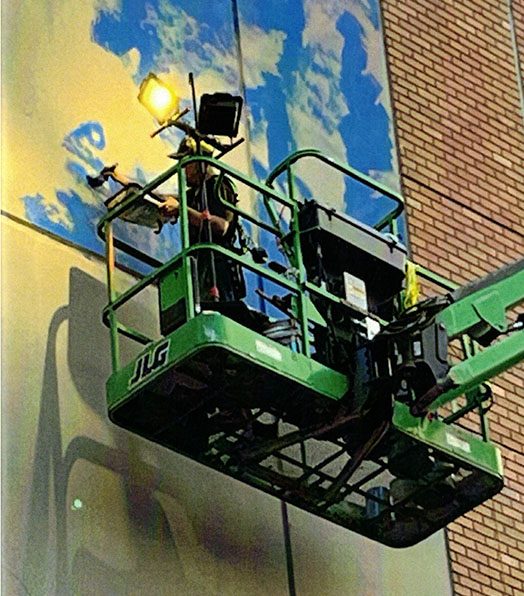 photo of artist Kevin Angulo standing in a cherry picker basket painting his mural