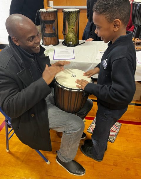 photo of a seated teaching artist holding an African drum and a standing student facing him. Both are playing the drum.