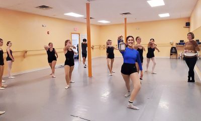 photo of a dance studio with numerous people in black leotards rehearsing. A single dancer is in blue, holding a lantern in the center of the frame.