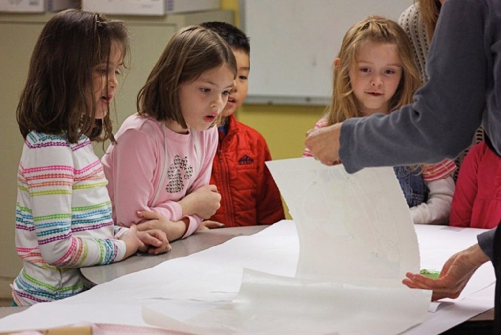 photo of 4 children standing at a table watching an adult pull back the paper to reveal a just-made print
