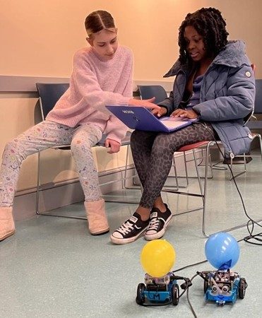 photo of 2 young people sitting, a couple of wheeled robots are on the floor in front of them. One youth has a laptop open while other youth is looking on, pointing at the computer screen