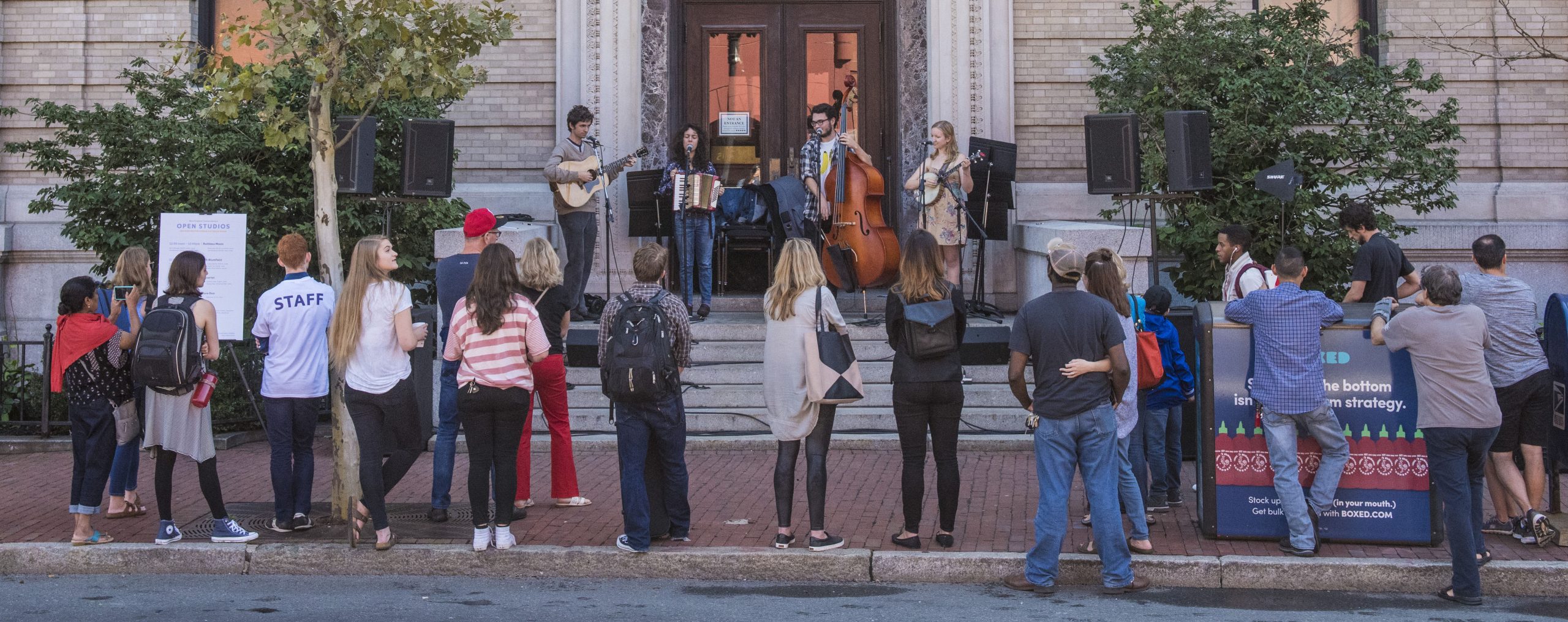 four musicians playing on the steps of a stone building, a line of spectators fills the sidewalk in from of them