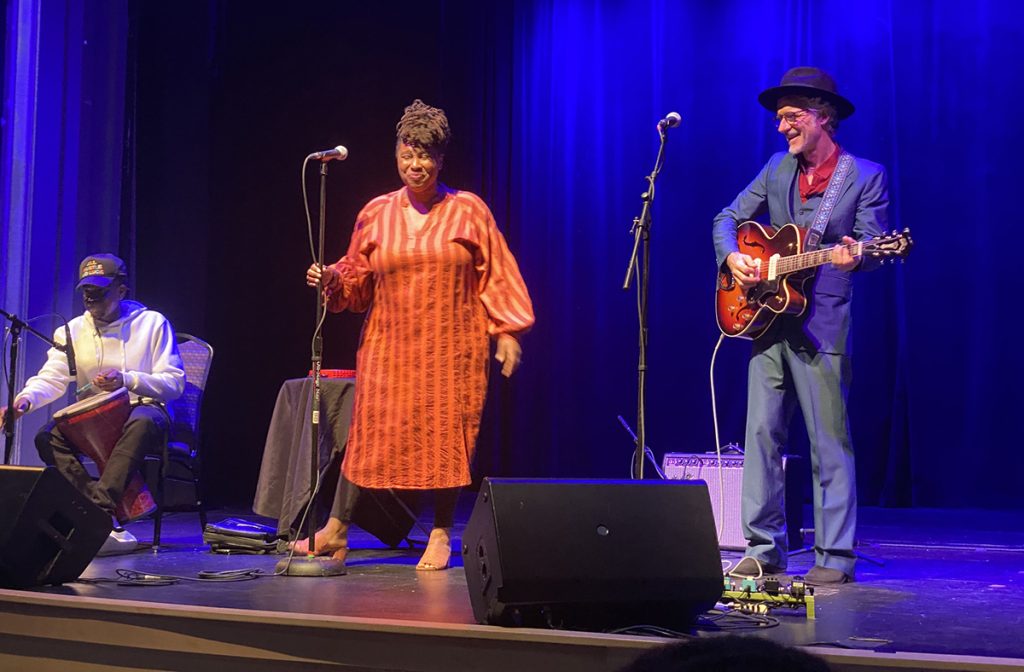 photo of 3 musicians performing on stage in front of a blue curtain backdrop. On the left a drummer in a baseball cap is seated in front of a mic. In the center the vocalist is wearing an orange dress over black legging. And on the right a smiling guitarist is wearing glasses and a wide brimmed hat.