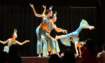 performance still of 4 dancers on a stage wearing traditional Chinese head pieces and colorful jumpsuits. The dancer on the right is doing a curved-back handstand