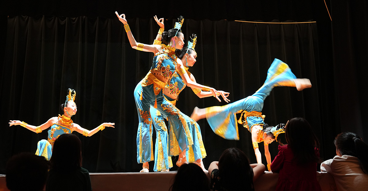 performance still of 4 dancers on a stage wearing traditional Chinese head pieces and colorful jumpsuits. The dancer on the right is doing a curved-back handstand