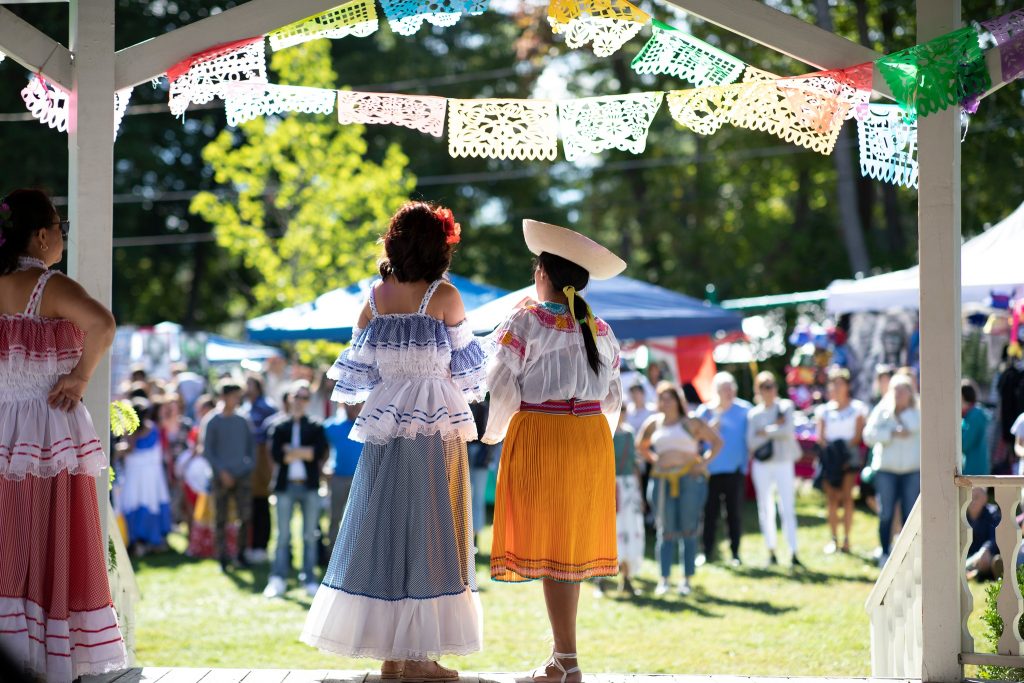 photo taken behind 2 performers on an outdoor stage, their backs to the camera. They are wearing traditional Latina skirts and tops, and are framed above by traditional Mexican paper flags. The background is a sunny green space with many people standing waiting for the show to start.