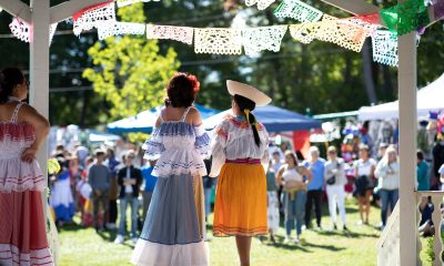 photo taken behind 2 performers on an outdoor stage, their backs to the camera. They are wearing traditional Latina skirts and tops, and are framed above by traditional Mexican paper flags. The background is a sunny green space with many people standing waiting for the show to start.