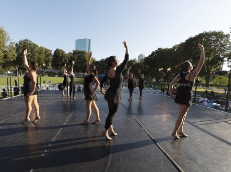 eight dancers in modern dance garb are positioned across an outdoor stage. In the background is the John Hancock Tower and a clear, blue sky.