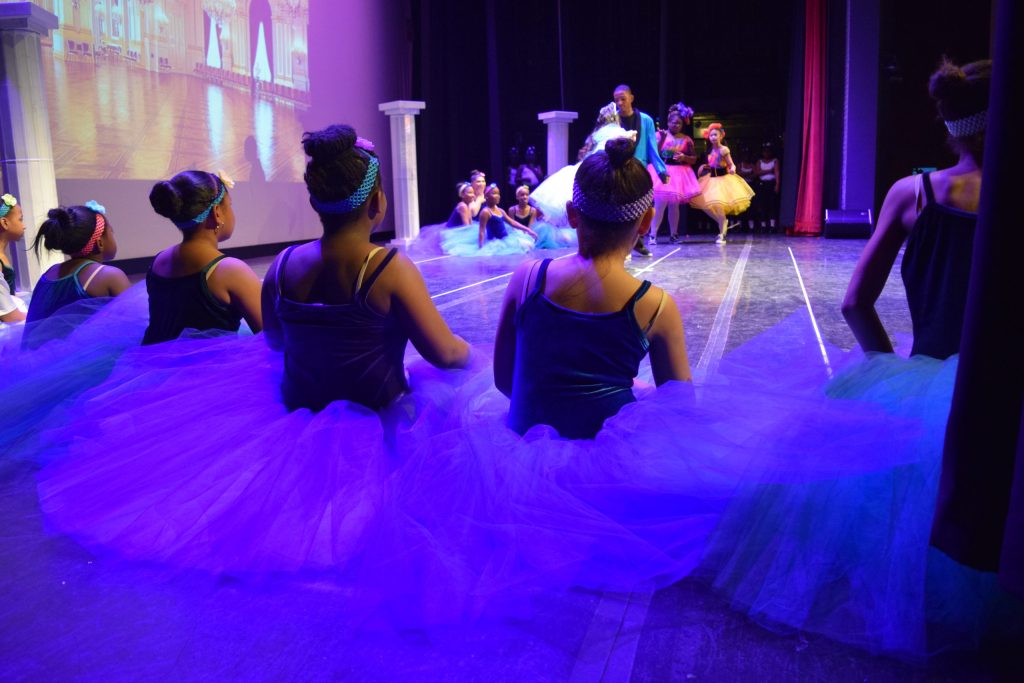 photo take from off stage. In the foreground 5 seated ballerinas look on at the main dancers performing. In the background the stage lights are focusing on a few other ballet dancers.