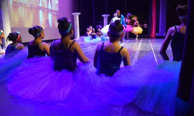 photo take from off stage. In the foreground 5 seated ballerinas are ready to go on for their part. In the background the stage lights are focusing on a few other ballet dancers.