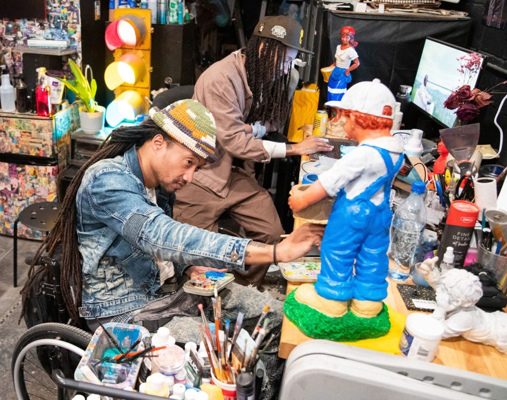 photo of two artists painting in their vibrant and cluttered studio. In the foreground, an artist sits in a wheelchair. In the background, another artist leans on a stool.