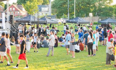 photo at twilight of people standing in a green field watching a performance, a row of black vendor tents is in the background.