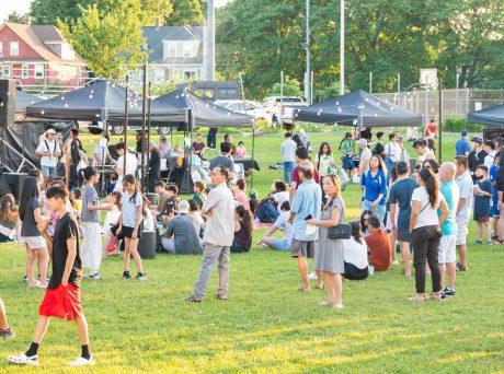 photo at twilight of people standing in a green field watching a performance, a row of black vendor tents is in the background.