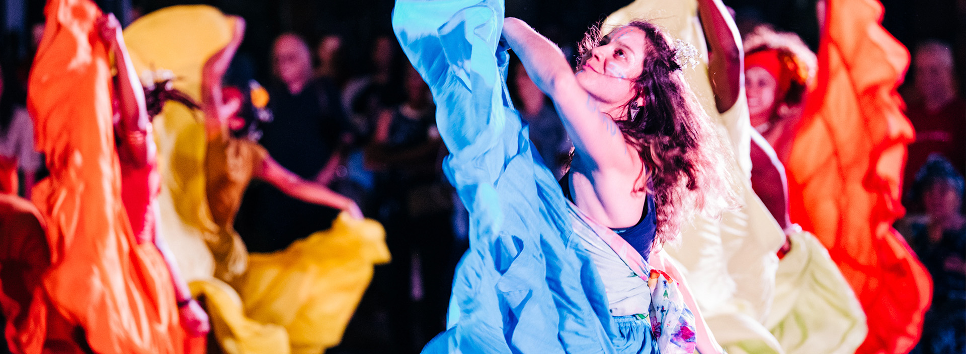 photo of an evening dance performance, a woman motions towards the sky while holding her blue flowing skirt, waves of fabric cascading down. Other similarly dressed dancers are on either side of her arms raised while holding their skirts. Their dresses are in oranges and yellows.