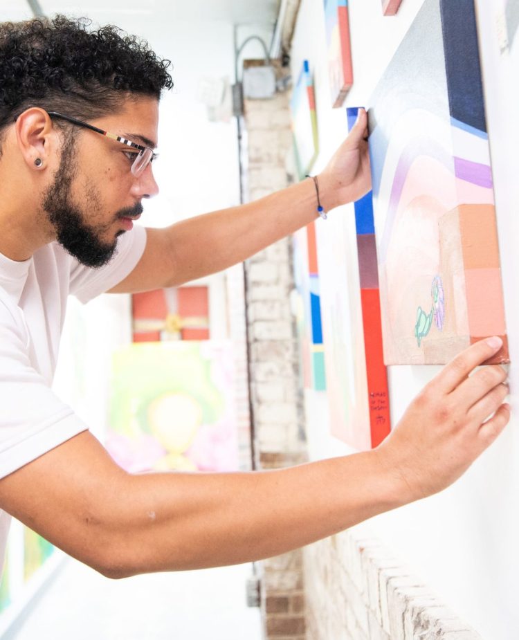 photo of an artist positioning one of his canvases along of wall covered with his canvases.