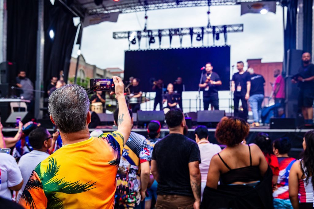 photo of a crowd watching an outdoor concert, the audiences' backs are facing the camera. In the foreground, a man wearing a bright orange tropical shirt holds up his phone to record the show.