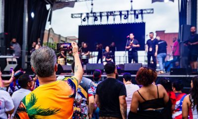 photo of a crowd watching an outdoor concert, the audiences' backs are facing the camera. In the foreground, a man wearing a bright orange tropical shirt holds up his phone to record the show.