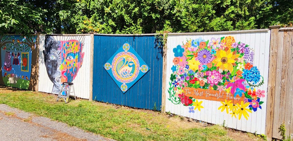 photo of 3 larger fence panels, each painted differently. one is a human face that's painted half in black and white and half in vibrant colors, next to that is a peacock blue panel with a diamond-shaped panel in its center. on that diamond is a painting of a peacock in a mosaic style. the last panel is covered almost entirely by a painting of a brilliant bouquet of flowers with the phrase the most beautiful flower underneath.